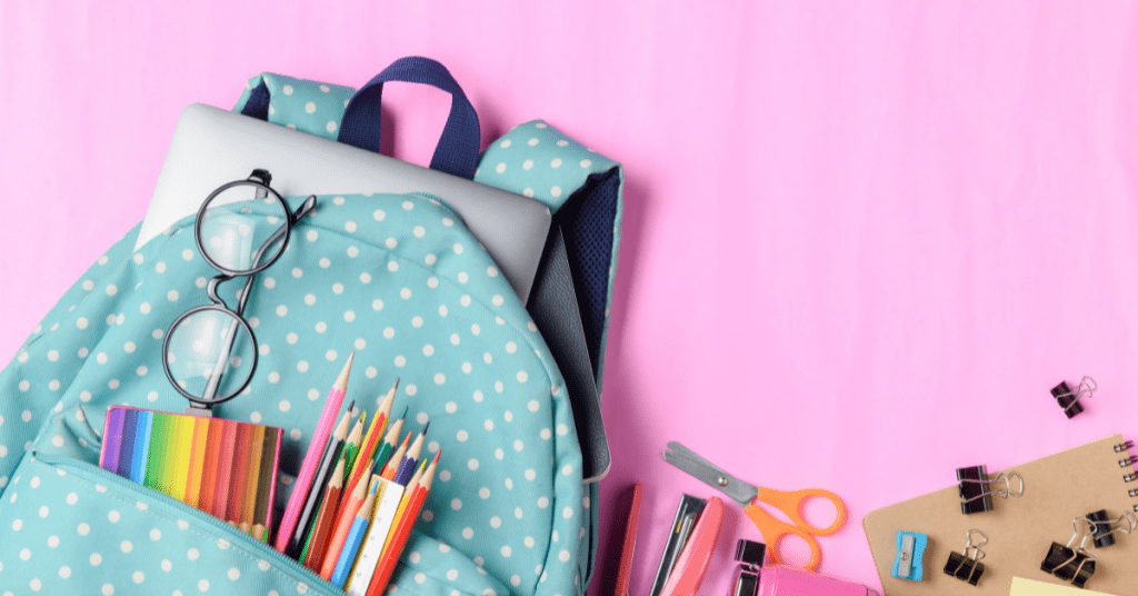 back to school items in book bag. Colored pencils, scissors, and pens on a pink background